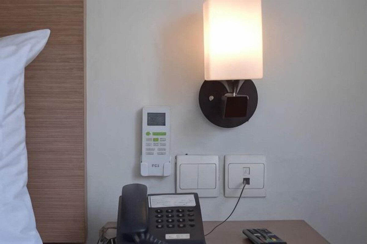 A hotel room with a bed, a bedside phone, a wall-mounted control panel, and a lamp.