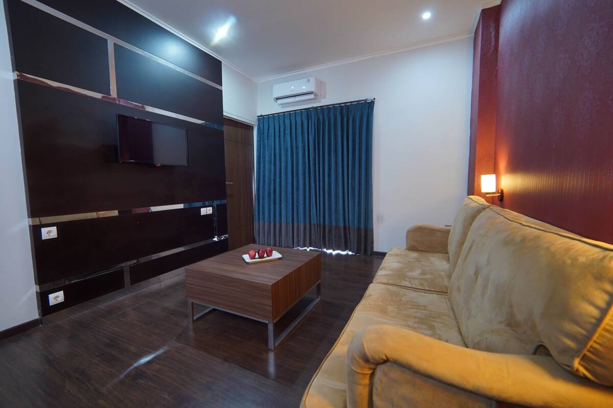 A modern living room with a flat-screen TV, a wooden coffee table, a beige sofa, and a blue curtain.