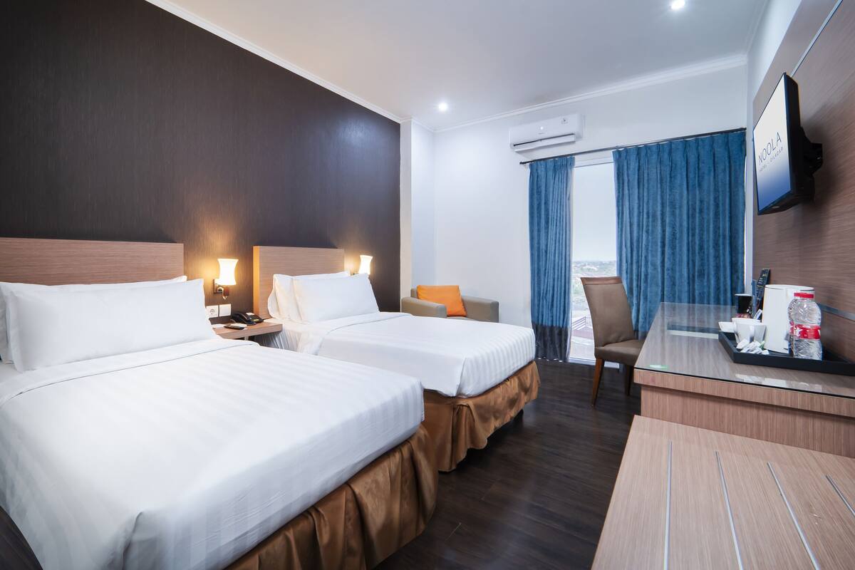 A hotel room with two beds, a desk, a chair, a TV, and a view of the city.