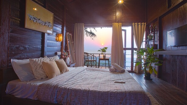 A wooden room with a bed, a small table with chairs, a potted plant, and a view of the sunset through a window. - Woodpegger By The Beach (Mandrem)