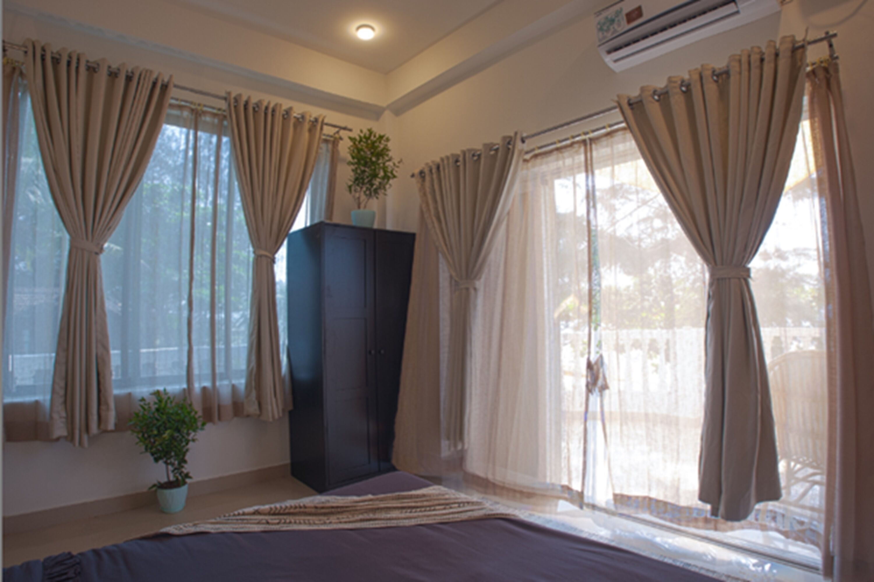 A bedroom with a bed, a potted plant, a dark-colored wardrobe, and large windows with curtains.