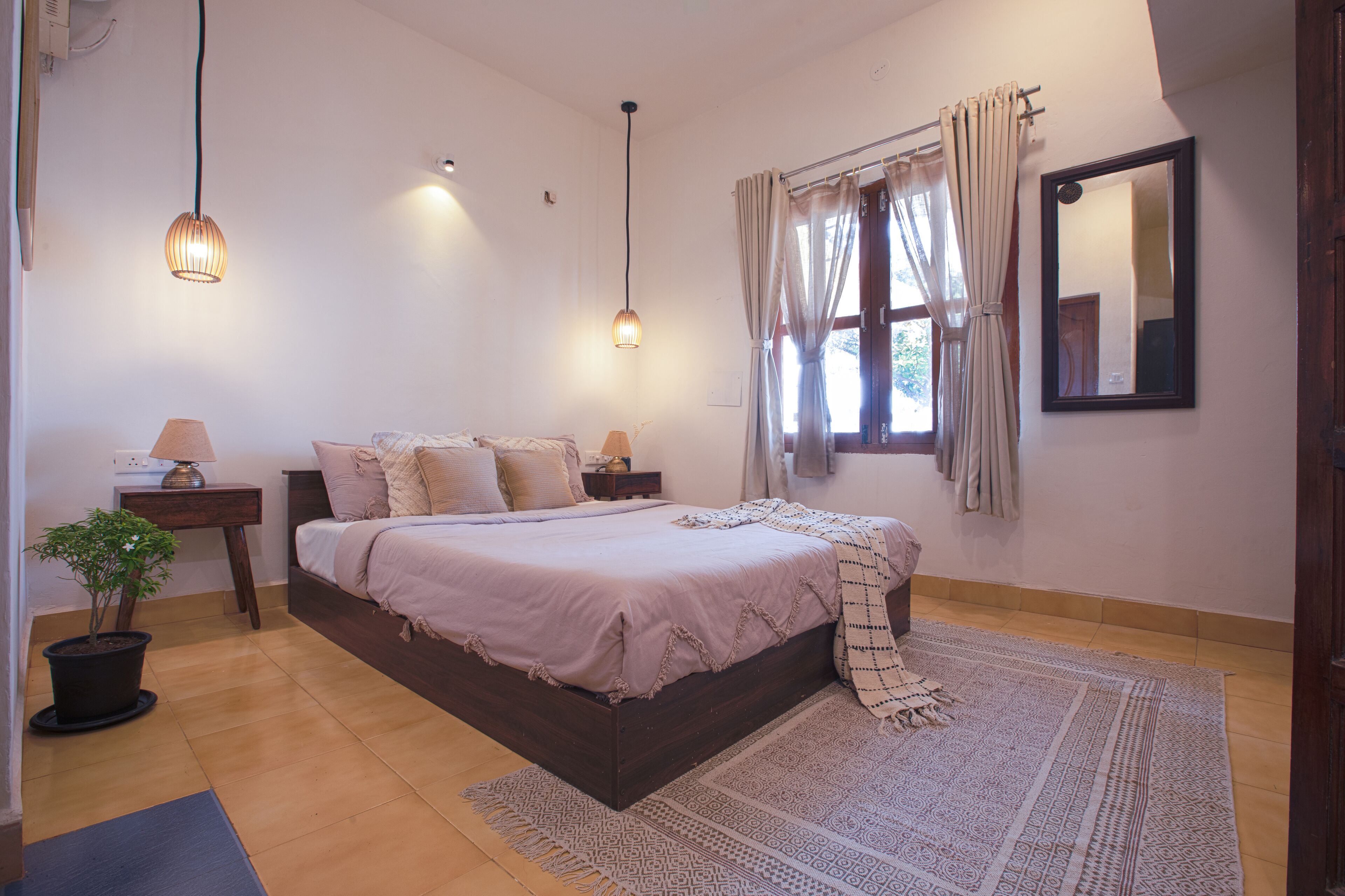 Une chambre à coucher avec un lit, des tables de chevet, une plante en pot, une fenêtre avec des rideaux et un miroir au mur.