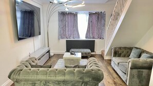 Un salon comprenant un grand téléviseur à écran plat, un canapé capitonné, un canapé plus petit et une table basse. Un escalier se trouve sur la droite, et il y a une fenêtre avec des rideaux.
