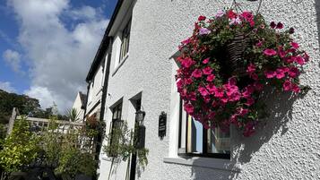 A white building with a hanging flower basket, a wooden table, and a blue chair outside.