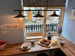 A breakfast setup with croissants, coffee, and juice on a wooden table, with a 'WELCOME' sign and a view of a street outside. - Beautiful 2-bed Cottage in Heysham Village (Morecambe)
