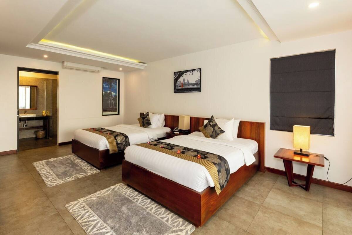 A hotel room with two beds, a bedside table with a lamp, a large window, and a painting on the wall.