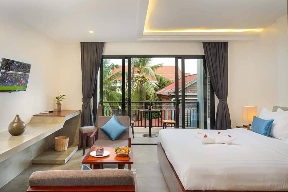 A hotel room with a bed, a sofa, a small table with a bowl of fruit, a TV on the wall, and a balcony with a view of palm trees and buildings.