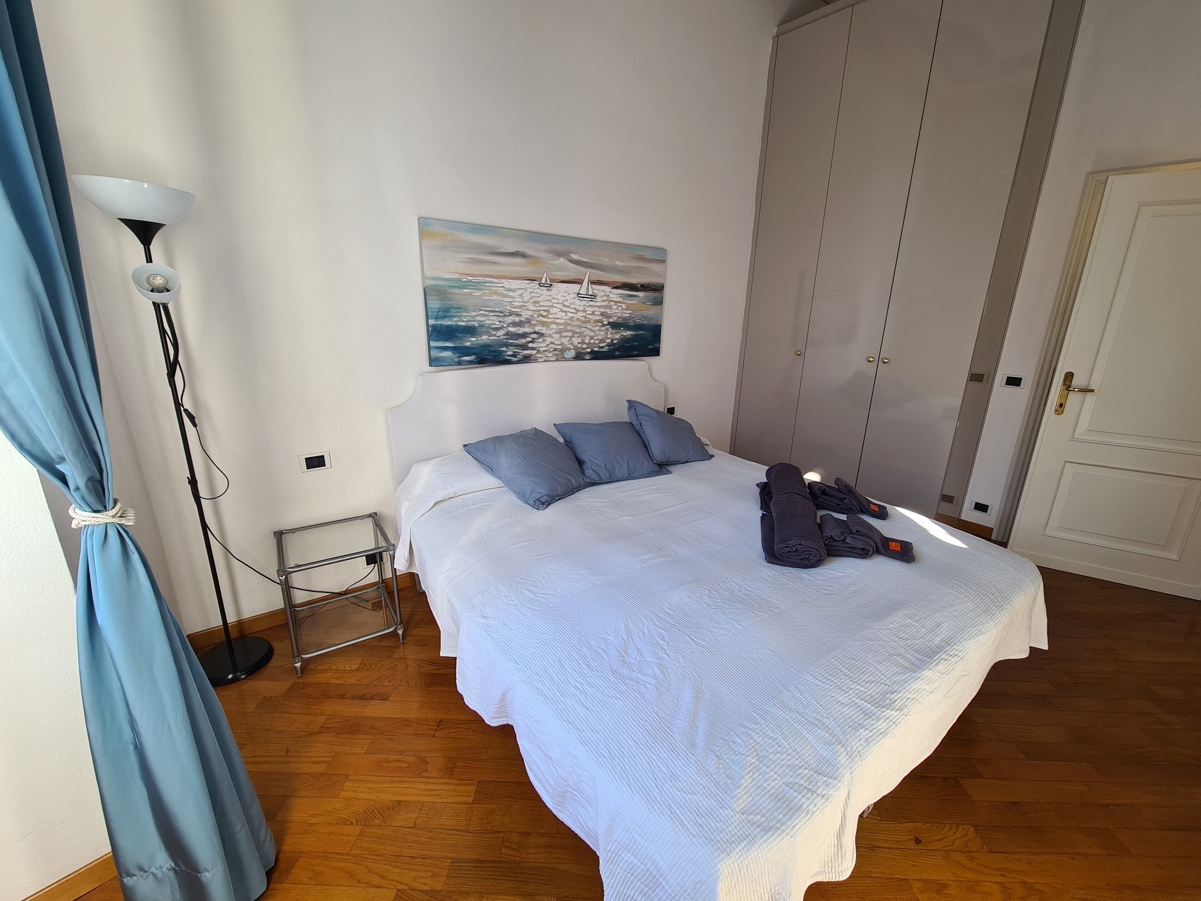 A bedroom with a bed, a painting of a seascape, a wardrobe, and a door.