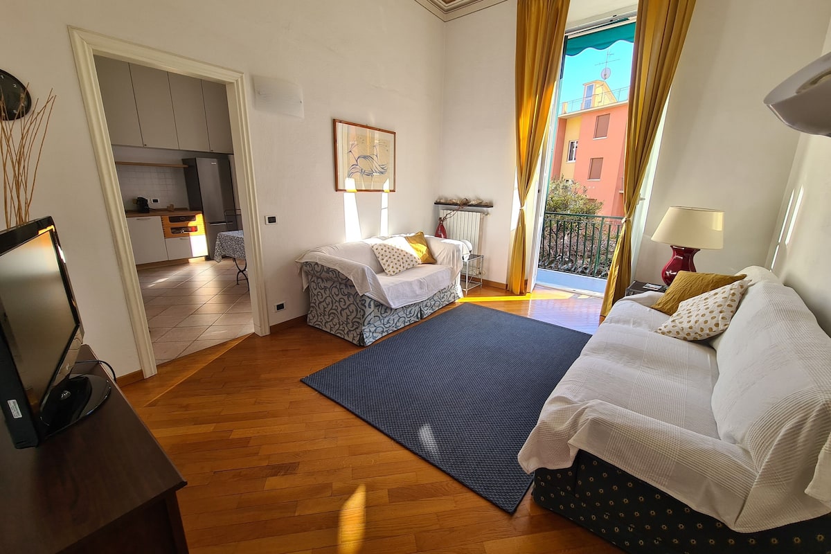 Habitación de hotel con dos camas, televisor, escritorio, zona de cocina y balcón con cortinas amarillas.
