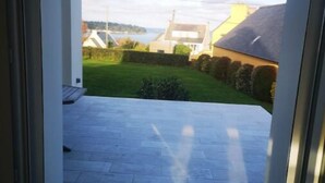 A view from a room's entrance to a patio area with a stone floor, a grassy lawn, and a building in the background. - Maison vue sur mer Avec Jardin (Carantec)