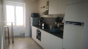 A compact kitchen with white cabinets, a microwave, an oven, and a sink. There is a window providing natural light and a small stool near the window. - Maison vue sur mer Avec Jardin (Carantec)
