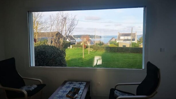 A room with a large window offering a view of a grassy area and buildings. There are two chairs and a small table with a book. - Maison vue sur mer Avec Jardin (Carantec)