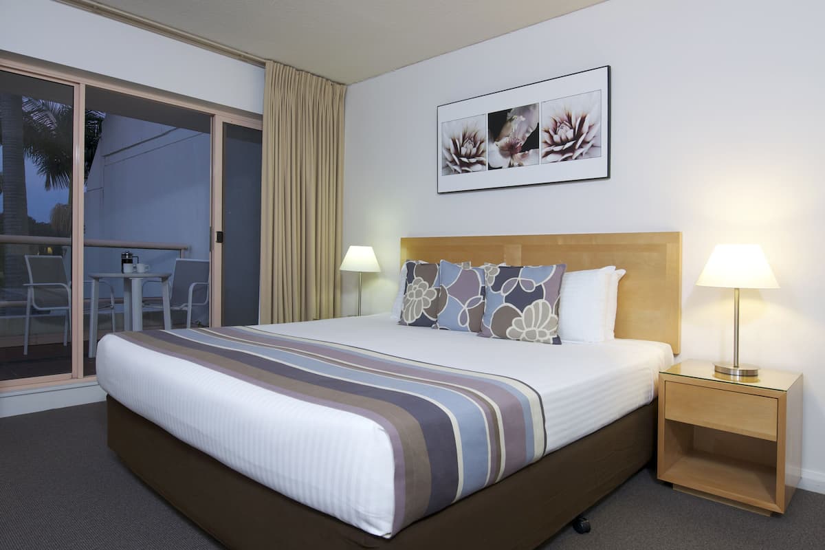 A hotel room with a large bed, two bedside lamps, a nightstand, a sliding glass door to a balcony, and a framed floral artwork on the wall.