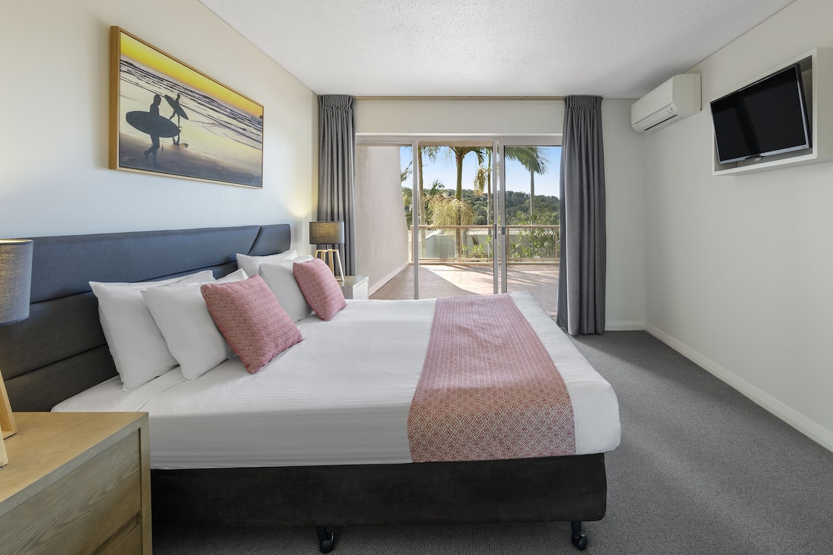 A hotel room with a large bed, a flat-screen TV, a painting of surfers, and a balcony with a view of palm trees.
