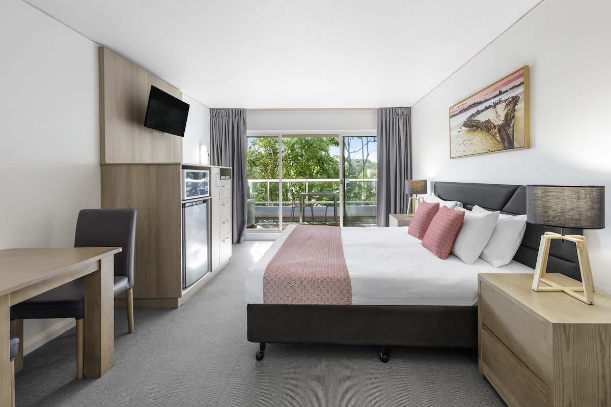 A hotel room with a bed, bedside table, TV, and a balcony with a view of trees.
