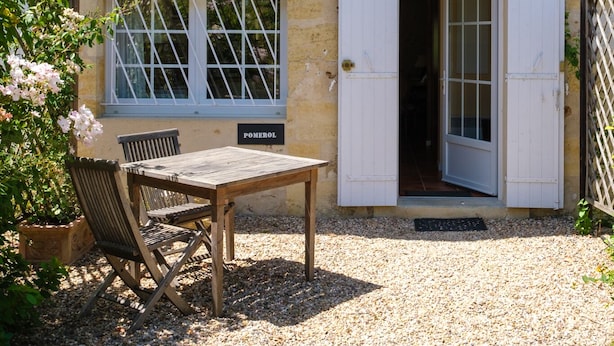 A rustic stone building with a wooden table and chairs outside, a door, and a window with shutters.