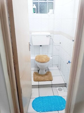 A small bathroom with a toilet, a blue mat on the floor, and a window with a bottle of lotion on the windowsill.
