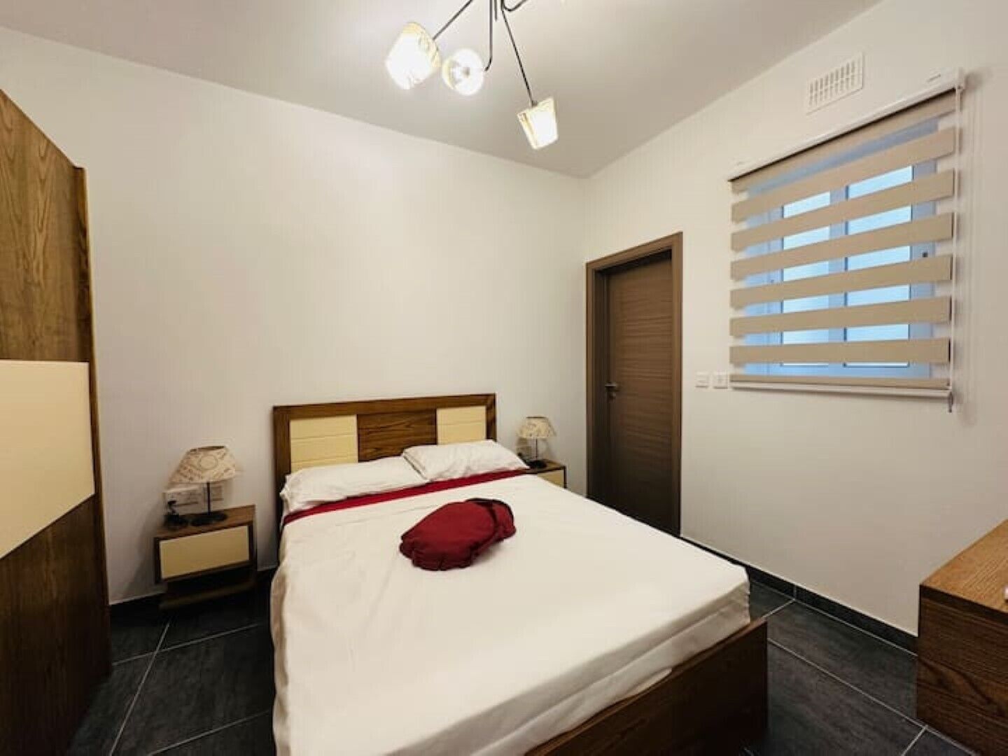 Un dormitorio con cama, mesitas de noche, un armario de madera, una ventana con persianas y una lámpara en el techo.