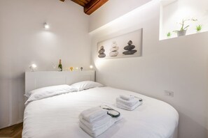 A neatly made bed with white linens, a bedside table with a bottle and glasses, and a wall-mounted artwork of stacked stones.