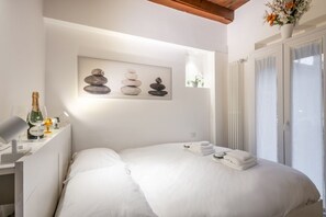 A hotel room with a bed, a desk with a bottle and glasses, a painting of stones, and a window with curtains.