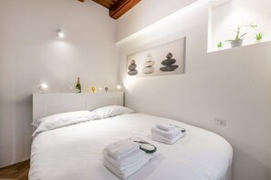 A neatly made bed with white linens, a bedside table with a bottle and glasses, and a wall-mounted artwork of stacked stones.