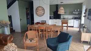 Uma sala ampla com uma mesa de jantar, cadeiras, uma poltrona azul e uma cozinha ao fundo.