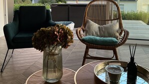 Uma sala de estar moderna com uma janela grande, uma cadeira de vime, um sofá escuro, um vaso de vidro com flores e um tabuleiro com uma vela.