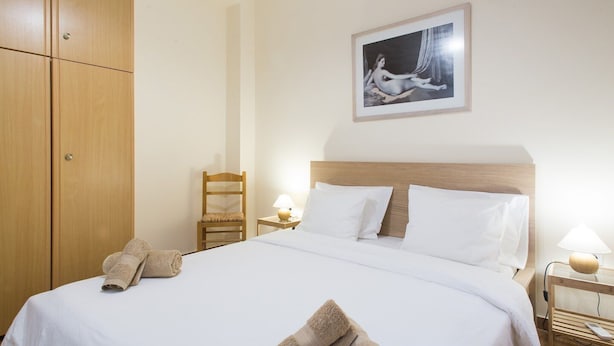 A hotel room with a bed, a wooden wardrobe, a chair, a nightstand with a lamp, and a framed picture on the wall.