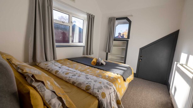 Une petite pièce bien éclairée, avec un lit, une fenêtre avec des rideaux, un radiateur et une porte.