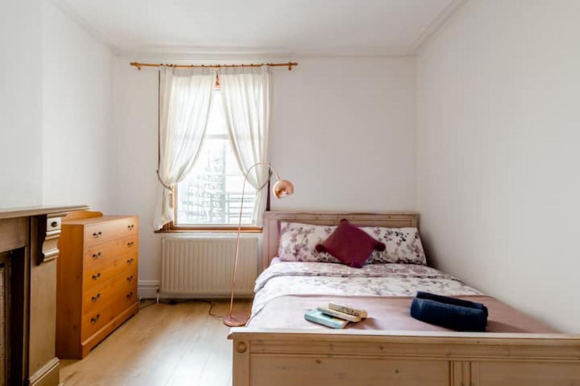 Un dormitorio con una cama de madera, una ventana con cortinas ligeras, un radiador y un aparador de madera.