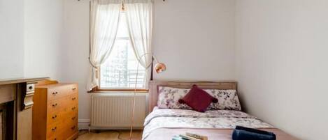 Un dormitorio con una cama de madera, una ventana con cortinas ligeras, un radiador y un aparador de madera.