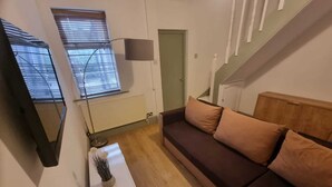 A living room with a brown sofa, a white side table, a floor lamp, and a window with blinds. - Elegant 4-bed House in Dartford (Dartford)