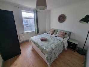 A bedroom with a bed, a radiator, a window with curtains, a bedside table, a lamp, and a wall decoration. - Elegant 4-bed House in Dartford (Dartford)