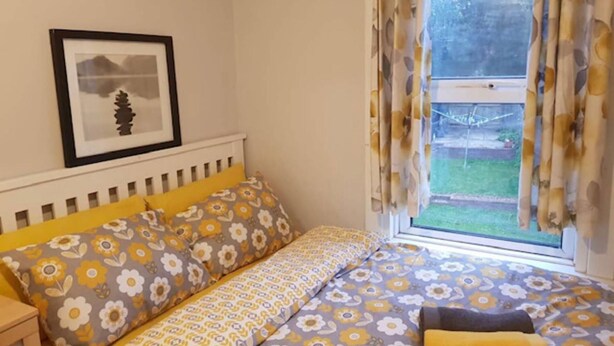A bedroom with a bed, a bedside table, a framed picture on the wall, and a window with curtains. - Gorgeous 4-bedroom House in Orpington (Orpington)