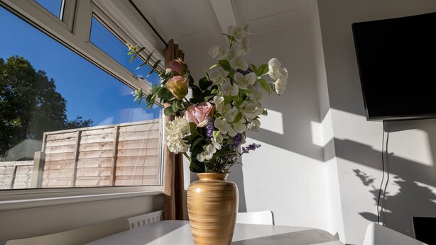A window with a wooden fence outside, a vase with flowers on a table, and a TV mounted on the wall. - Immaculate 2-bed Apartment in Dartford (Dartford)
