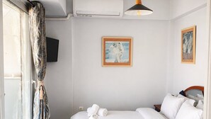 A hotel room with a bed, a television, a painting, and an air conditioning unit.