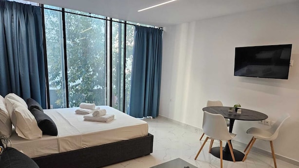 A modern hotel room with a bed, a sofa, a dining table with chairs, and a TV mounted on the wall.