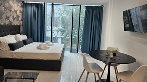A modern bedroom with a large bed, a TV, a dining table with chairs, and a view of the outdoors.