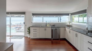 A modern kitchen with white cabinets, a stainless steel dishwasher, and a large window overlooking a pool. - View Point: Ocean Views & Near CBD (Burnie)