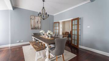 A dining room with a dark wooden table, white chairs, a chandelier, and a painting on the wall.