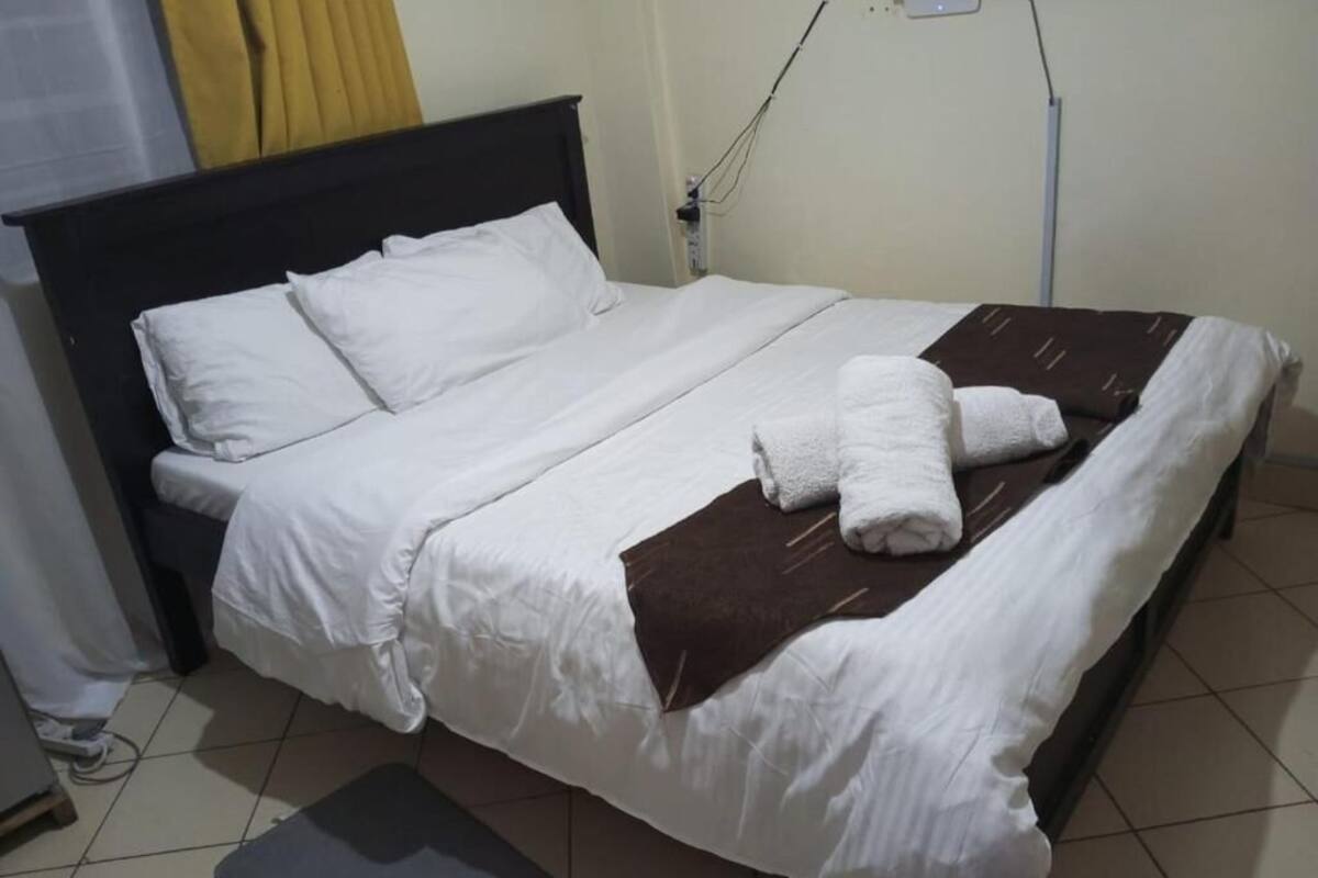 A neatly made bed with white linens and a brown blanket, a gray mat at the foot of the bed, and a visible electrical outlet on the wall.