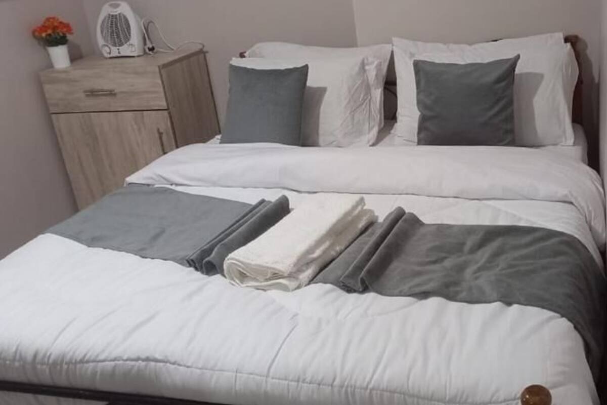 A neatly made bed with white and gray bedding, a wooden nightstand with a small plant, and a white wall.