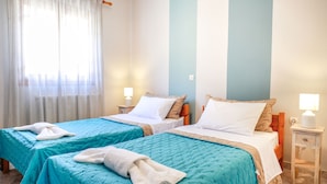 Habitación de hotel con dos camas, cada una con cubrecama azul y almohadas blancas. Se observa un mesita de noche con lámpara y cajón.