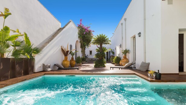 A swimming pool with a wooden deck, surrounded by white walls and potted plants.