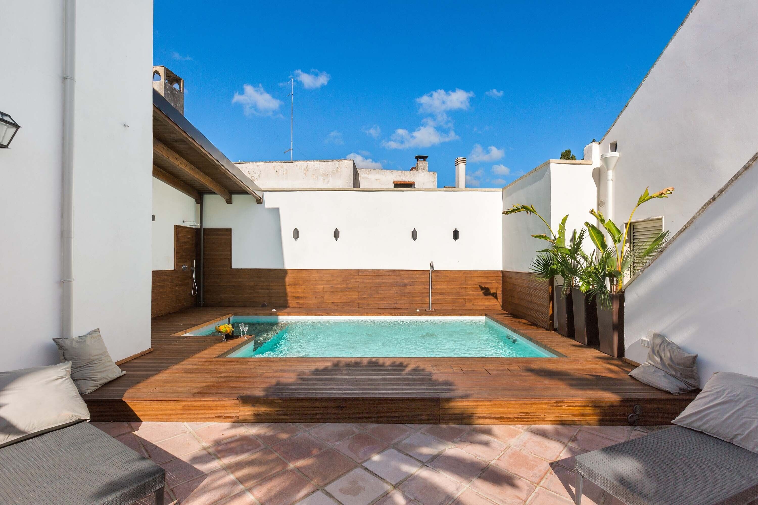 A rooftop pool with wooden decking and a small jacuzzi, surrounded by white walls and a clear blue sky.