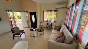 A spacious living room with a beige sofa, a white chair, and a small table with decorative items. There is a door with a glass panel, a window with sheer curtains, and an air conditioning unit on the wall.