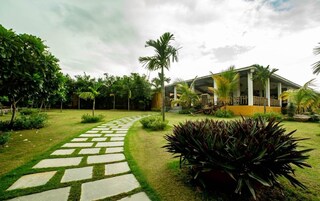Um edifício moderno com área externa coberta, rodeado por vegetação bem cuidada e um caminho de pedra que leva à entrada.