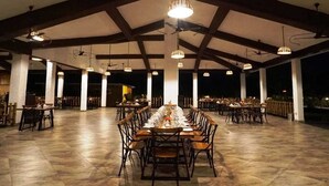 An outdoor dining area with a long table set for a meal, surrounded by chairs, under a high ceiling with exposed beams.