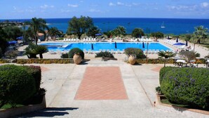 A resort pool area with clear blue water, surrounded by lounge chairs and umbrellas, with a view of the ocean and palm trees. - Villaggio Club Dolomiti Sul Mare (Briatico)