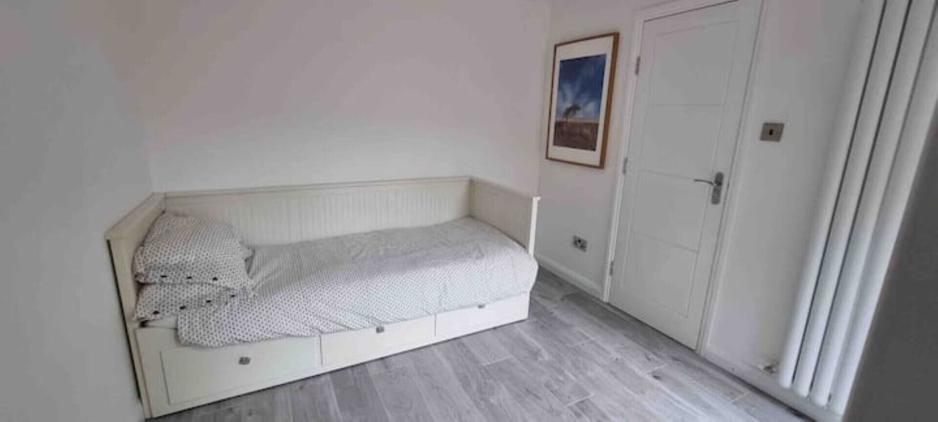 Une petite chambre avec un lit simple, un lit blanc, une porte et un tableau encadré au mur.